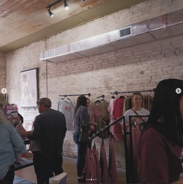 A wide shot of some shirts and bookshelves against a pale pink wall with people shopping