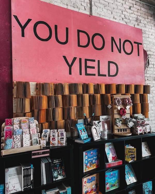 A pink sign that says YOU DO NOT YIELD with paperback stacked in an open fanned position below atop some shelves with books, bookmarks and stickers 