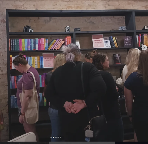 A woman with grey hair in a ponytail stands with her hands folded behind her back looking at shelf of contemporary romances while other people are shopping 