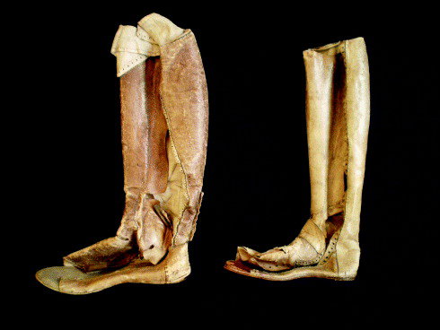 Side view of extremely old, worn to tatters boot liners that wrap mostly around the calf, and inside one has a higher heel base to compensate for height. There is also padding at one of the calves