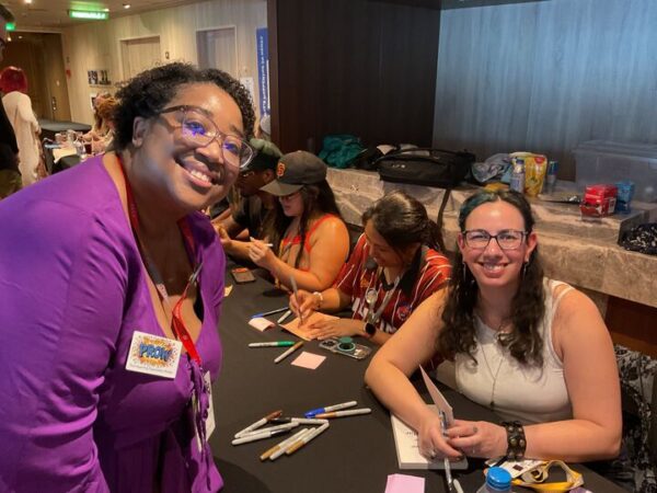Shana, a Black woman in a purple top with short curly hair, leans toward Amal El-Motar, who is seated. She has long wavy hair and glasses. they're both smiling at the camera