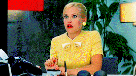 A blonde woman in a yellow dress with a bow at the neck faints away from her desk. As she falls her heels, also yellow, appear on the desk