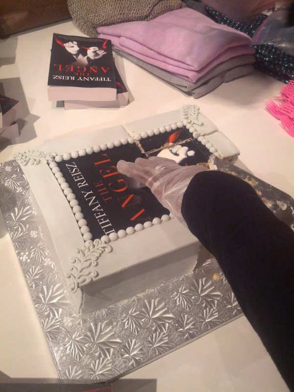 A gloved hand cutting a cake with an image of Tiffany Reisz's book The Angel