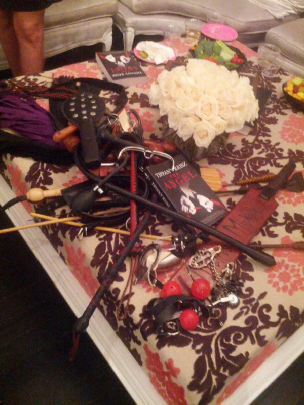 A pile of domination sex toys including whips, paddles, and restraints, alongside a bouquet of white roses on a patterned ottoman 