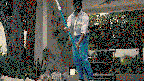 A man in blue pants, sneakers, and a blue 90s shirt is slowly skimming a pool