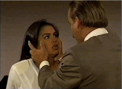 A woman with brown skin and dark hair is being held by the face by a guy in a suit. The camera zooms in over his shoulder as he shakes her and then she passes out