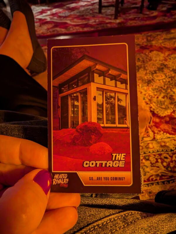 A photo of my left hand holding a playing card with a picture of The Cottage from the show, and the caption The Cottage. Below that: are you coming...?