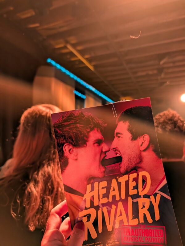 my heated rivalry musical program, a single card with a photograph of the two leads biting a hockey puck facing each other and grimacing, with the stage behind the program.