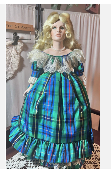 A close up of the plaid blonde doll from an eBay listing. The porcelain face has wide eyes and peach lips, a pearl necklace, and a voluminous empire waist plaid gown. 