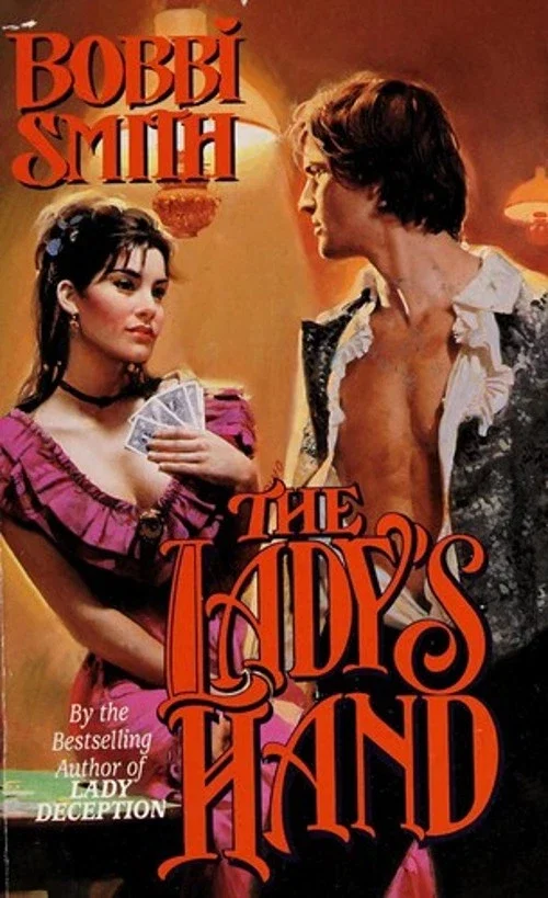 Bobbi Smith’s The Lady’s Hand A white woman with dark hair in a half updo and bangs is lounging on a green topped table. She is wearing a dark purple ruffled gown with a wide neck and short sleeves, and she’s holding a full hand of cars against her chest. In front of her is a man with tousled hair, a brocade vest and a ruffly shirt, with the shirt wide open AND STILL TUCKED IN ofc, and he’s semi-facing the reader looking over his shoulder at the woman. 