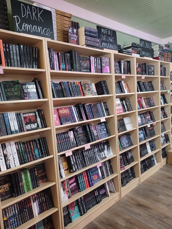 A picture of full bookshelves that look to be tilted backwards so the front edge is higher than the back, allowing the spines to be visible on all shelves. At the top are blackboards saying Dark Romance, Mafia/Outlaws Romance, and Why Choose Romance.