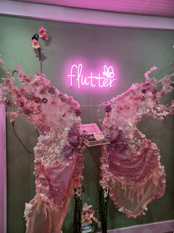 An installation of giant pink wings in flowers, gauze, chiffon and other diaphanous fabrics, with a jewelry box in the middle and a neon sign that reads Flutter in pink script 