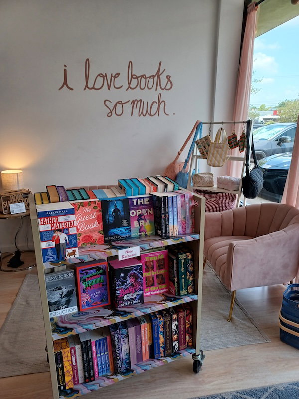 The cart of new releases at Finney's against a wall that reads I LOVE BOOK SO MUCH. Behind the cart you can see beige comfy chairs and a display of soft handbags 