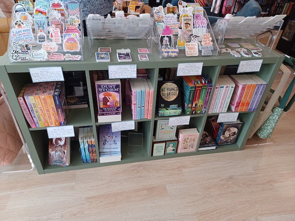 A green-grey set of dight cubbies with books, shelf talkers, and special editions, and on the top is a display of stickers of all shapes and sizes 