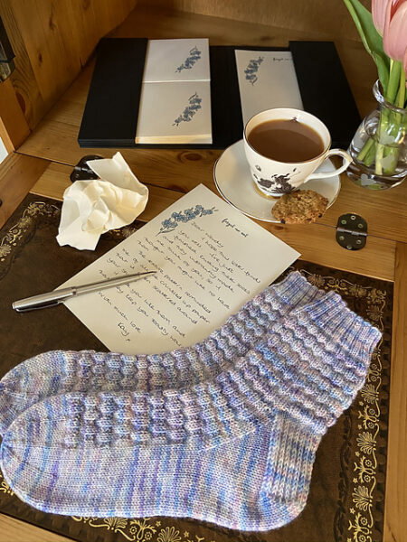 A pair of blue and purple textured socks sit on a table with a handwritten letter and a cup of tea