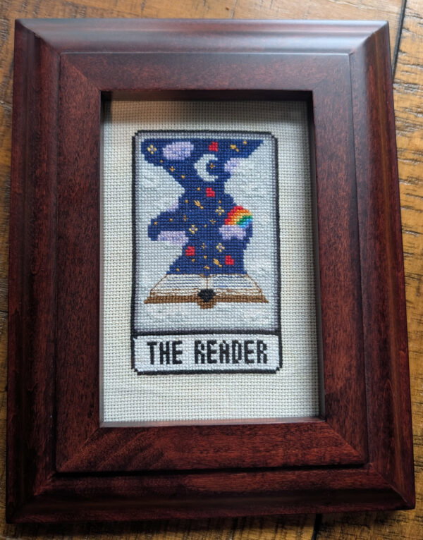 A 4.5" x 2.5" hand stitched tarot card for "The Reader" - clearly not a major arcana but it is now! This embroidery piece is mounted in a 9x7 frame that can be hung or displayed on a shelf. The card depicts an open book with stars, clouds, rainbows and hearts and is a design created by The Colourful Coven UK.