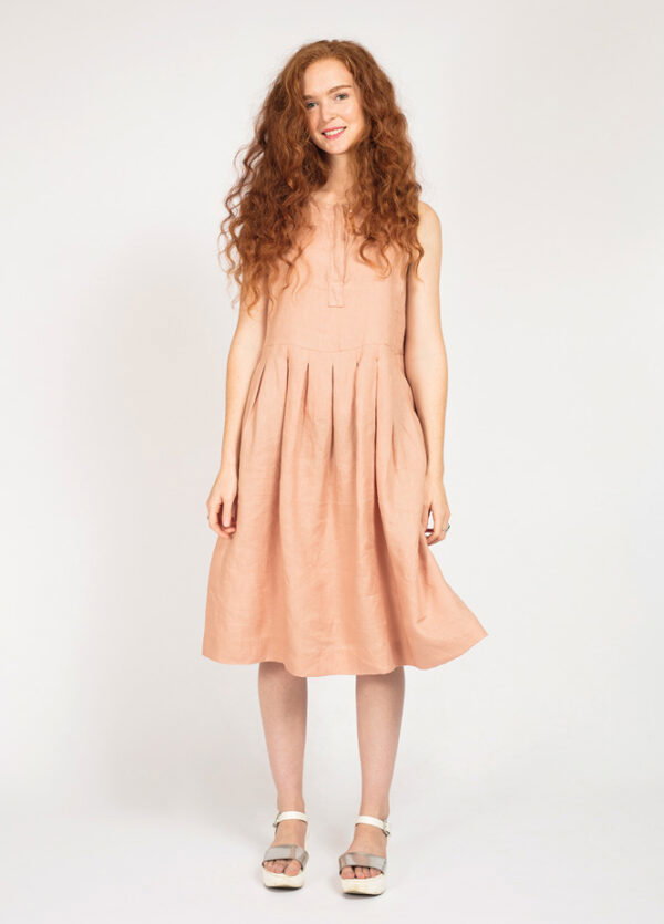 a white woman with red curly hair is standing wearing a peach colored dress with a pleated front and a tie neck. No sleeves. the dress is just below her knees 