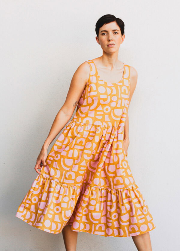 A white woman with a short dark pixie cut wearing a sleeveless dress with two ruffled tiers in a orange and pink geometric print fabric. The dress comes down below her knees 