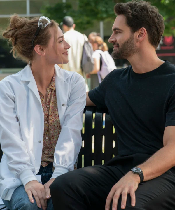 Lili Reinhart and Tom Bateman facing each other on a bench. Lili has her hair up, and is smiling at him, and she's wearing a white lab coat. Tom has dark hair, a beard, and a black tshirt on